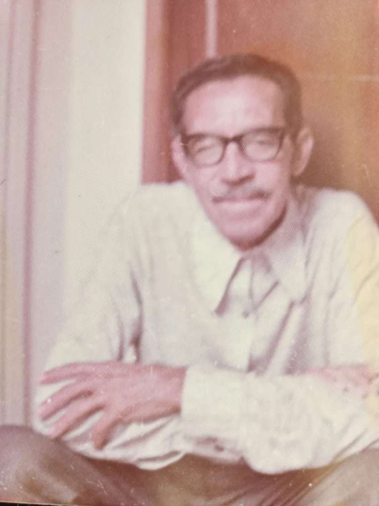 Foto antiga e desbotada de um senhor de óculos e bigode, com os braços cruzados sobre a mesa: o avô do autor