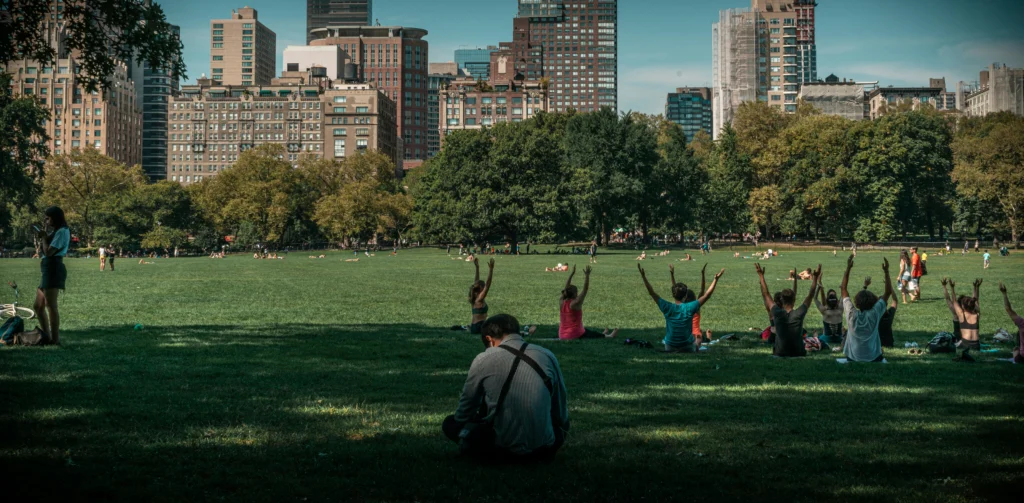 Pessoas sentadas na grama de um parque, com os braços levantados, fazendo yoga; prédios ao fundo