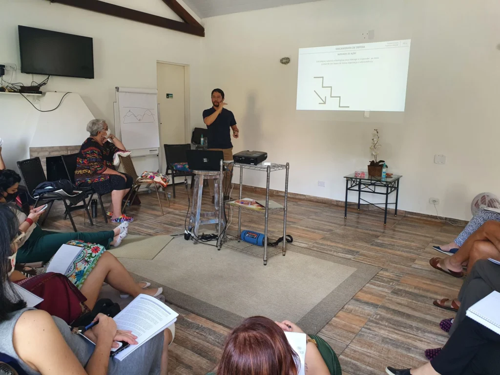Francisco Masuda dando uma palestra para um grupo sentado em roda, em uma sala ampla, com um slide projetado na parede