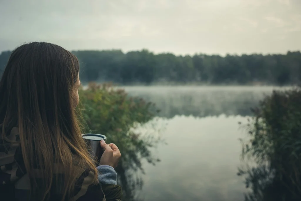 Mulher de costas, com uma xícara na mão, diante de um lago com neblina